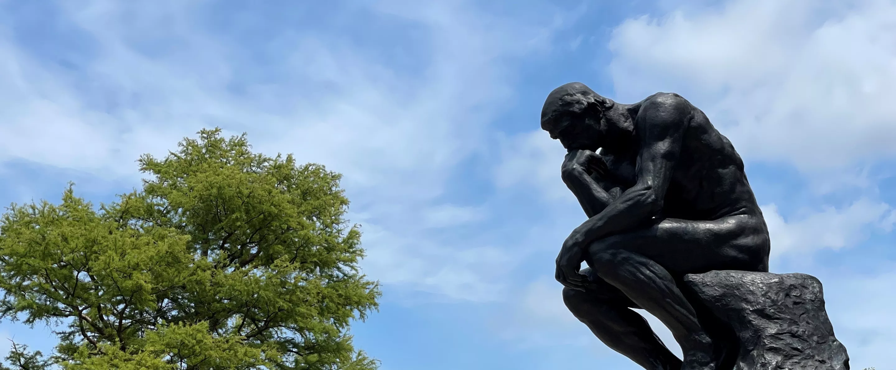 UofL's thinker statue from the side with the sky in the background. Trees on campus can be seen in the background as well as a lamp post with a UofL banner.