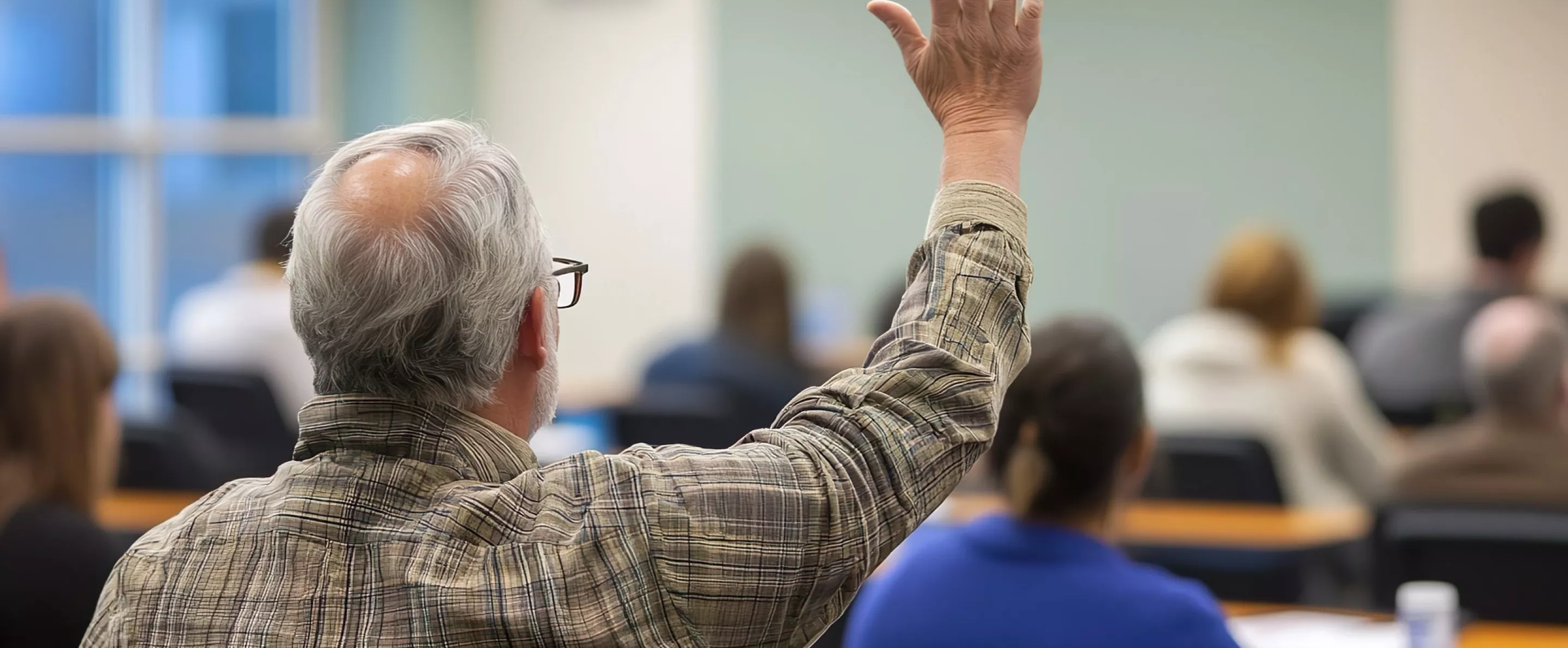 Back view of older student raising his hand to answer teacher's question during education training class.