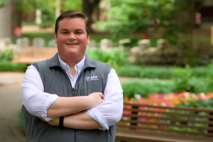 Person smiling with crossed arms, wearing a white shirt and grey vest with "AFP KENTUCKY" logo, standing in an outdoor, leafy setting.