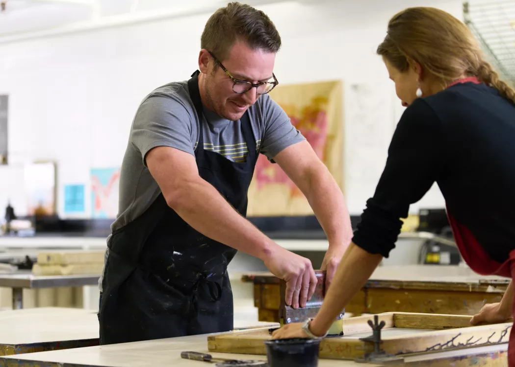 Student pulling a screen print with Rachel Singel, Associate Professor in a printmaking studio course