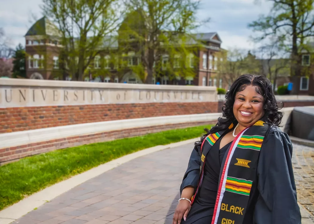 Young woman in graduation gown and kente stole sitting by a wall with "UNIVERSITY of LOUISVILLE" inscribed on it.