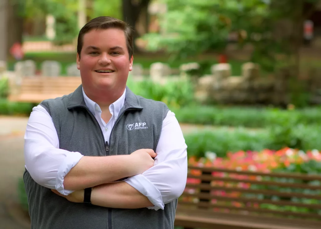 Person smiling with crossed arms, wearing a white shirt and grey vest with "AFP KENTUCKY" logo, standing in an outdoor, leafy setting.