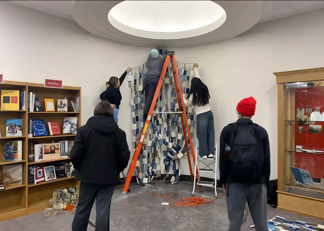 A group of individuals collaborates on a large fabric project inside a library setting, surrounded by books and resources.