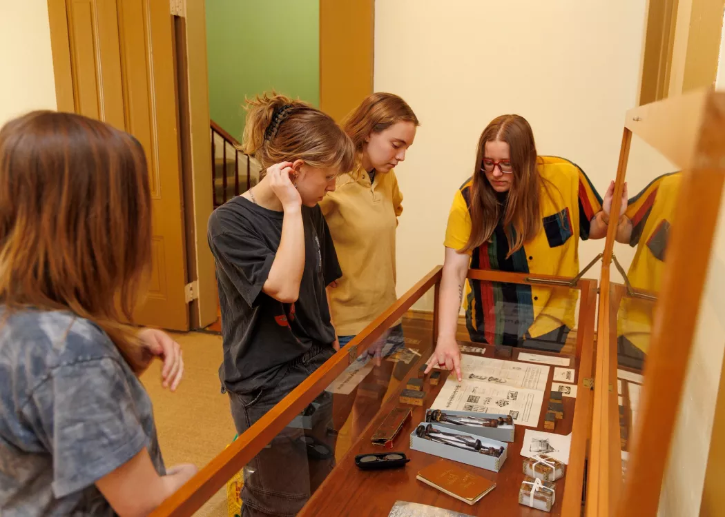 Lewis leans over a wood-rimmed vitrine, teaching their research interns about Lee H. Stephens’ engraving tools, wood…