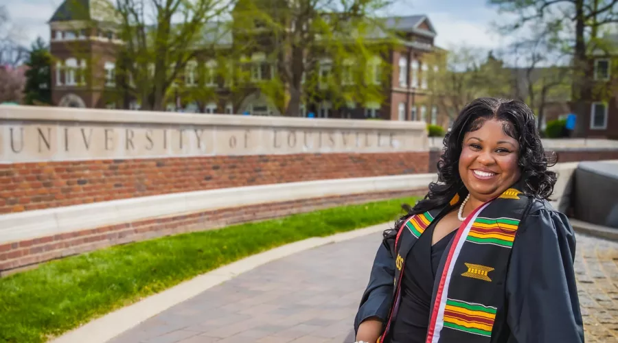 Young woman in graduation gown and kente stole sitting by a wall with "UNIVERSITY of LOUISVILLE" inscribed on it.