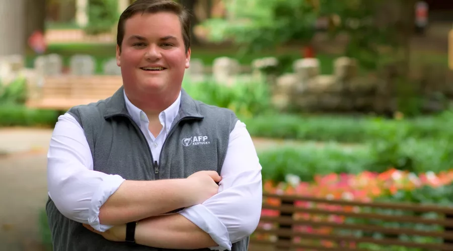 Person smiling with crossed arms, wearing a white shirt and grey vest with "AFP KENTUCKY" logo, standing in an outdoor, leafy setting.
