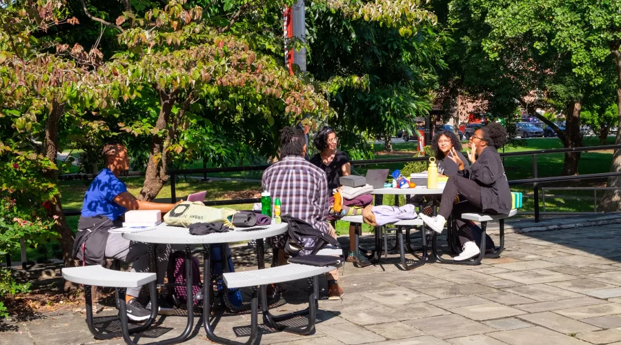 Group out students of color sitting at two picnic tables outside of Schneider building.